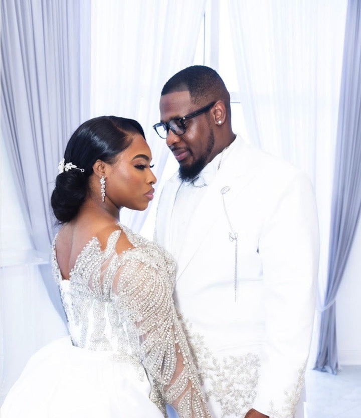 Man and woman in wedding attire standing close together against a white curtain.